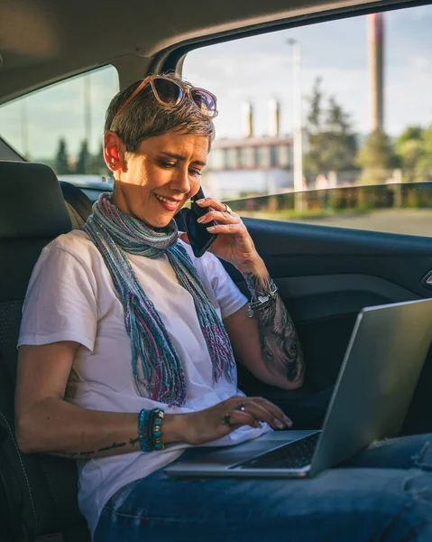 ラップトップコンピュータで作業している車の後部座席に座っている1人の成熟した女性白人女性は カジュアルコピースペースを身に着けている道路上の短い灰色の髪の現代と夏の日に電話で話をする — ストック写真