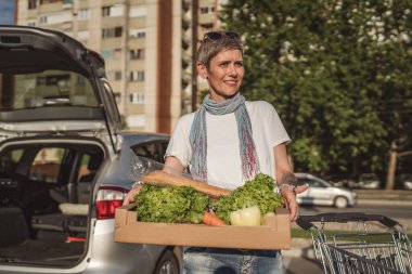 Yetişkin bir kadın, otoparkta duran beyaz bir kadın süpermarketin önünde elinde yiyecek ve sebzelerle duruyor. Mutlu bir gülümseme, sağlıklı vegan konsepti yiyor.