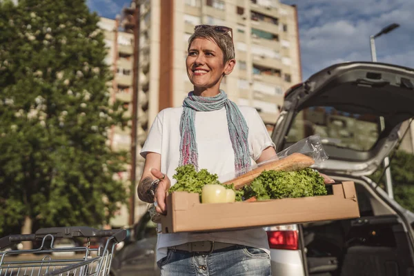 One woman mature caucasian female standing in the parking lot bu car in front of supermarket grocery store holding box with food and vegetables in sunny day happy smile healthy eating vegan concept