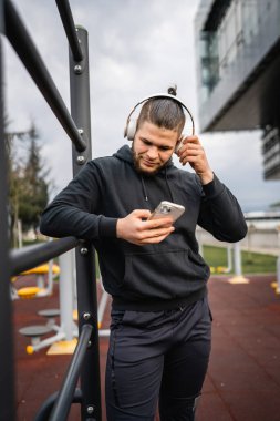 Beyaz tenli genç bir erkek açık hava eğitim parkı spor salonunda duruyor. Kulaklıklı cep telefonu kullanıyor. Gerçek insanları eğitmek için internetten mesajlar yolluyor.