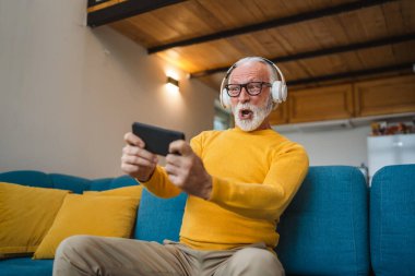 Yaşlı bir beyaz adam evinde mutlu bir şekilde oturur video oyunları oynar, eğlenir cep telefonu bulundurur, akıllı telefon alır ve eğlenceli fotokopi çekerler.