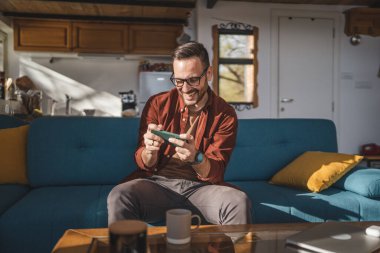 Yetişkin bir beyaz adam evinde oturup mutlu bir şekilde gülümser video oyunları oynar, eğlenir cep telefonu tutar, akıllı telefon tutar, fotokopi çeker gözlük takar ve gömlek giyer.