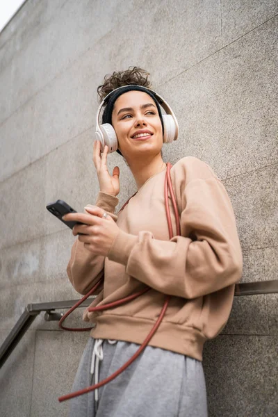 One woman young adult generation z caucasian modern female with headphones and skipping rope happy smile use mobile phone app for training or music browse online in the city real person copy space