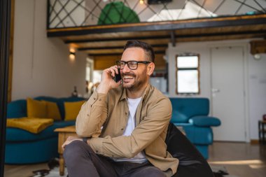 Yetişkin bir beyaz erkek cep telefonuyla arama yapar. Mutlu bir gülümseme ile konuşur. Evde kanepede oturur, gömlek giyer ve gözlük takar.