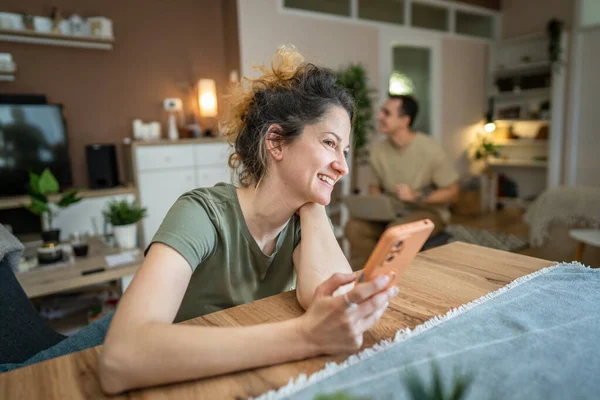 一人の女性は Smsメッセージのために自宅でスマートフォンを使用するか バックグラウンドで彼女の夫と一緒にテーブルに座ってインターネットを参照してください現代的な技術家族生活の概念コピースペースリアル人 — ストック写真