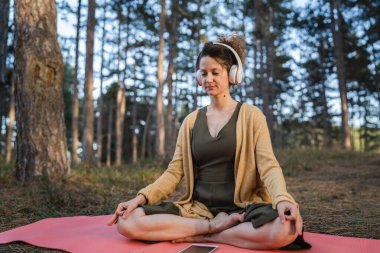 Yetişkin bir kadın, parkta ya da ormanda tek başına oturmuş elinde kulaklık tutarak meditasyona hazırlanıyor. Kişisel bakım konsepti.