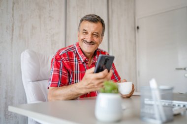 Üst düzey bir erkek girişimci iş yerindeki masada oturuyor bir fincan kahve ve cep telefonu tutuyor online mesajları veya sosyal medya içeriğini kontrol ediyor gerçek insanlar boşluğu kopyalıyor
