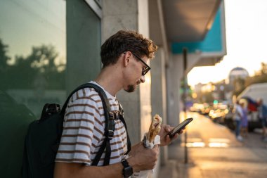 Genç, yetişkin, beyaz bir erkek, güneşli bir günde şehirde sandviç yiyor, fast food konsepti yiyor, şehir yaşamının fotokopisini çekiyor, turistleri duvarın önünde yiyor, gerçek bir insan cep telefonu kullanıyor.
