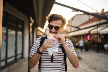 Gündüz vakti şehirde genç, yetişkin, beyaz bir erkek sandviç yiyor. Fast food konsepti. Şehir hayatı.