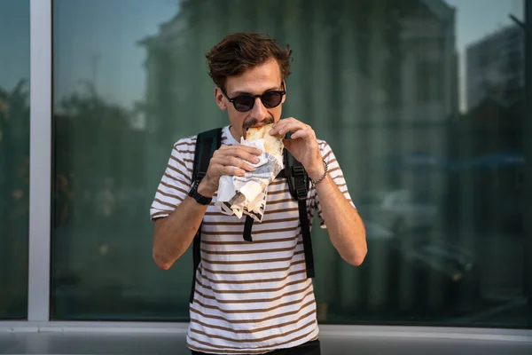 晴れた日のスタンドで市内の1人の若い大人の現代的な白人男性とサンドイッチを食べますファーストフードのコンセプト都市生活コピースペース観光客は壁の前で食べる本物の人 — ストック写真