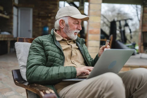 一位高年级的白种人 留着胡子 整天坐在户外用笔记本电脑在网上购物 他买东西时 手里拿着信用卡 享受着自己的退休生活 — 图库照片