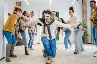 Yetişkin bir beyaz erkek, bir grup erkek ve kadın arkadaşın önünde sanal gerçeklik cihazının keyfini çıkarıyor. Takım oluşturma semineri sırasında birlikte eğleniyorlar. Gerçek insanlar parlak filtreler.