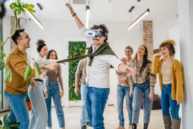 Yetişkin bir beyaz erkek, bir grup erkek ve kadın arkadaşın önünde sanal gerçeklik cihazının keyfini çıkarıyor. Takım oluşturma semineri sırasında birlikte eğleniyorlar. Gerçek insanlar parlak filtreler.
