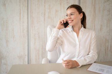 One woman female caucasian entrepreneur businesswoman or secretary sitting at her office at desk work happy smile making a phone call talk checking paper documents contract wear white shirt copy space
