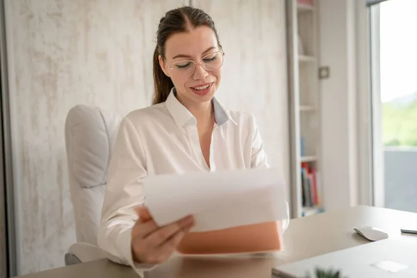 Stock image One woman female caucasian entrepreneur businesswoman or secretary sitting at her office at desk work happy smile checking paper documents contract wear white shirt copy space