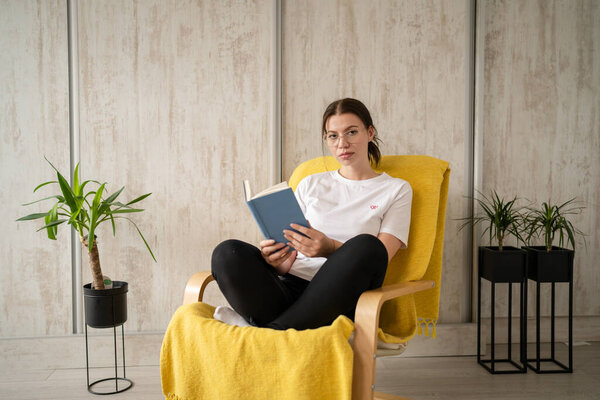 Одна женщина, молодая белая женщина сидит в кресле дома читать книгу копия пространства передний вид реальных людей досуг концепции выходные глядя на камеру серьезно