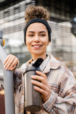 Kulaklıklı bir genç kadın eğitim için hazırlanıyor. Su içmek için destek şişesi tutuyor. Açık havada güzel beyaz kadın nesli.
