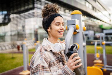 Kulaklıklı bir genç kadın eğitim için hazırlanıyor. Su içmek için destek şişesi tutuyor. Açık havada güzel beyaz kadın nesli.