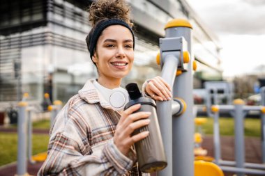 Kulaklıklı bir genç kadın eğitim için hazırlanıyor. Su içmek için destek şişesi tutuyor. Açık havada güzel beyaz kadın nesli.