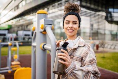 Kulaklıklı bir genç kadın eğitim için hazırlanıyor. Su içmek için destek şişesi tutuyor. Açık havada güzel beyaz kadın nesli.