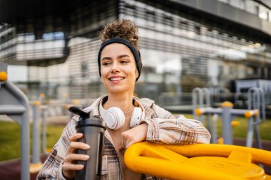 Kulaklıklı bir genç kadın eğitim için hazırlanıyor. Su içmek için destek şişesi tutuyor. Açık havada güzel beyaz kadın nesli.