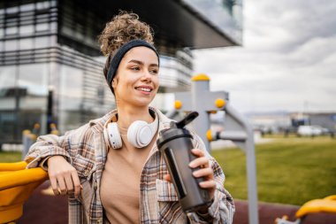 Kulaklıklı bir genç kadın eğitim için hazırlanıyor. Su içmek için destek şişesi tutuyor. Açık havada güzel beyaz kadın nesli.