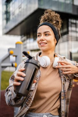 Kulaklıklı bir genç kadın eğitim için hazırlanıyor. Su içmek için destek şişesi tutuyor. Açık havada güzel beyaz kadın nesli.