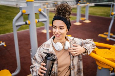 Kulaklıklı bir genç kadın eğitim için hazırlanıyor. Su içmek için destek şişesi tutuyor. Açık havada güzel beyaz kadın nesli.