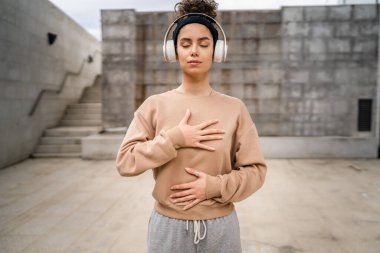 Yetişkin beyaz bir kadın internet rehberli meditasyon için kulaklık kullanıyor. Gözü kapalı farkındalık tezahürü uyguluyor. Açık havada duruyor.