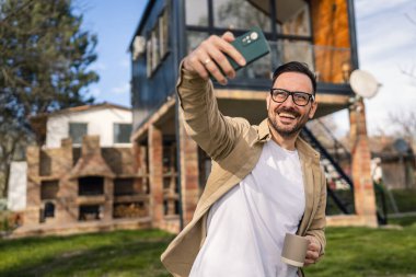 Kafkasyalı mutlu bir erkek, tatilde gerçek insanlar uzay boşluğunu kopyalarken modern küçük bir evin önünde selfie ya da video çağrısı için akıllı telefon kullanır.