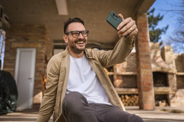 Kafkasyalı mutlu bir erkek, tatilde gerçek insanlar uzay boşluğunu kopyalarken modern küçük bir evin önünde selfie ya da video çağrısı için akıllı telefon kullanır.