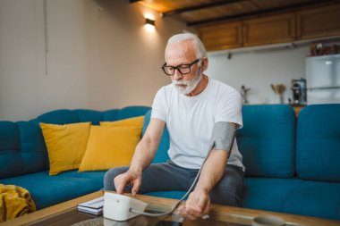 Sakallı yaşlı beyaz bir erkek evde tek başına otururken kan basıncı ölçme cihazı kullanıyor. Gerçek insanlar uzay sağlığı konseptini kopyalıyor.