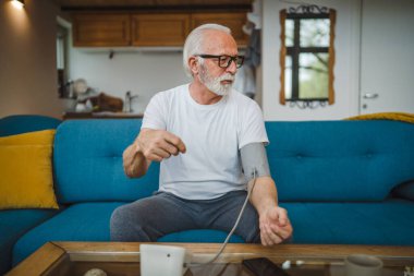 Sakallı yaşlı beyaz bir erkek evde tek başına otururken kan basıncı ölçme cihazı kullanıyor. Gerçek insanlar uzay sağlığı konseptini kopyalıyor.