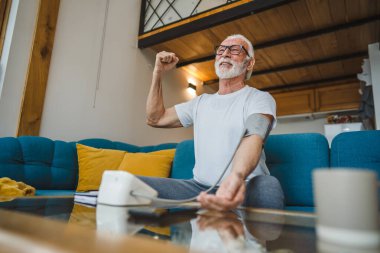 Mutlu, yaşlı, sakallı, sağlıklı beyaz bir erkek, kan basıncı cihazını kullanarak evde tek başına oturup sonuçları ölçüyor. Gerçek insanlar uzay sağlık hizmetleri konseptini kopyalıyor.