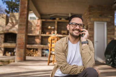 Sakallı ve gözlüklü yetişkin bir adam güneşli bir günde dışarıda oturur mutlu bir şekilde gülümser cep telefonunu kullanarak konuşma yeri fotokopisi çektirir gerçek insanlar gibi.