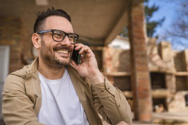 Sakallı ve gözlüklü yetişkin bir adam güneşli bir günde dışarıda oturur mutlu bir şekilde gülümser cep telefonunu kullanarak konuşma yeri fotokopisi çektirir gerçek insanlar gibi.