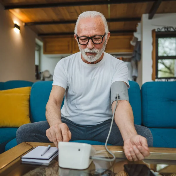 stock image One man senior caucasian male with beard use blood pressure device to check and measure results while sit at the table at home alone real people copy space health care concept