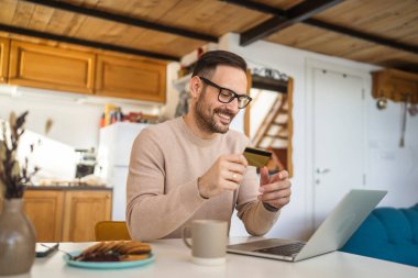 Yetişkin bir beyaz erkek evde dizüstü bilgisayarla mutlu bir gülümseme içinde banka kredi kartı alışverişi yapıyor gerçek insanlar e-ticaret e-bankacılık hizmetlerini kopyalıyor.