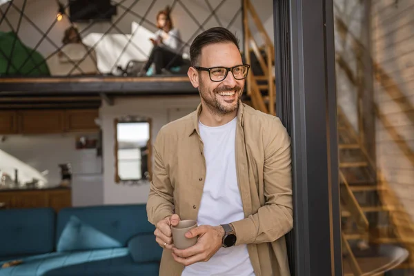 一个留着胡子和眼镜的成年高加索男子站在门口的阳台上 开心地微笑着 端着一杯咖啡 模仿空间每天早晨的日常概念 — 图库照片