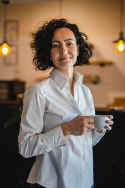Bir kadın evdeki ya da işyerindeki pencerenin önünde duruyor. Girişimci kadın beyaz gömlek giyiyor. Kahve içiyor, gülümsüyor.