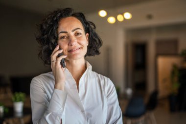 One woman mature caucasian female businesswoman entrepreneur stand at work or home use mobile phone making a call talk real people copy space wear white shirt curly hair happy smile