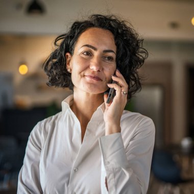 One woman mature caucasian female businesswoman entrepreneur stand at work or home use mobile phone making a call talk real people copy space wear white shirt curly hair happy smile