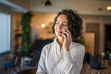 Yetişkin bir kadın Kafkasyalı iş kadını iş yerinde ya da ev telefonuyla konuşuyor. Gerçek insanlar konuşma yapıyor. Gerçek insanlar beyaz gömlek giyiyor, kıvırcık saçlar takıyor.