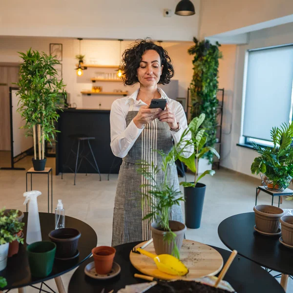 One woman mature adult caucasian female take care of her plants at home photographing flower pot with her smartphone mobile phone send photos to social media or as message real person