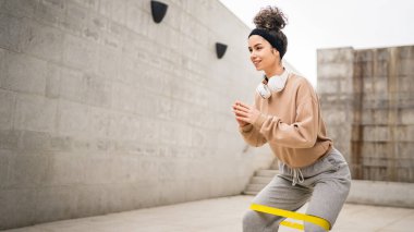 Bir yetişkin beyaz kadın sporcu şehrin dışında esmer ve sağlıklı esmer kadınlar için esneme konseptinde esnemiş beton arka planda elastik lastik direnç bantları ile eğitim görüyor.