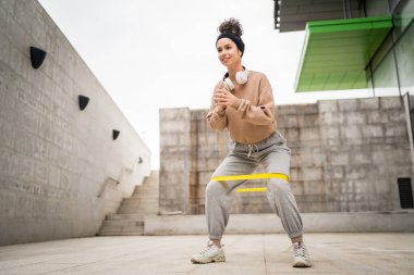 Bir yetişkin beyaz kadın sporcu şehrin dışında esmer ve sağlıklı esmer kadınlar için esneme konseptinde esnemiş beton arka planda elastik lastik direnç bantları ile eğitim görüyor.