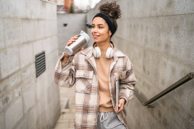 Genç bir kadın yetişkin kız öğrenci ayakta duruyor ya da dışarıda yürüyor dizüstü bilgisayar ve bir fincan kahve tutuyor gerçek bir insan uzayı kopyalıyor mutlu bir gülümseme kulaklıkla gün içinde kendine güveniyor.