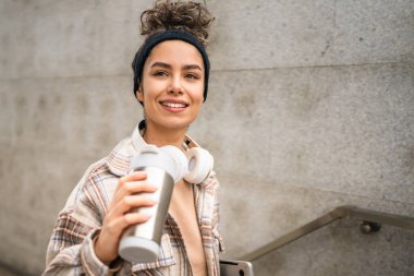 Genç bir kadın yetişkin kız öğrenci ayakta duruyor ya da dışarıda yürüyor dizüstü bilgisayar ve bir fincan kahve tutuyor gerçek bir insan uzayı kopyalıyor mutlu bir gülümseme kulaklıkla gün içinde kendine güveniyor.