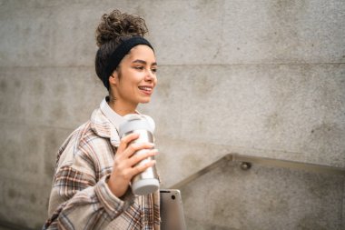 Genç bir kadın yetişkin kız öğrenci ayakta duruyor ya da dışarıda yürüyor dizüstü bilgisayar ve bir fincan kahve tutuyor gerçek bir insan uzayı kopyalıyor mutlu bir gülümseme kulaklıkla gün içinde kendine güveniyor.