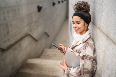 Genç bir kadın yetişkin kız öğrenci ayakta duruyor ya da dışarıda geziyor dizüstü bilgisayarı kullanıyor SMS mesajları için akıllı telefonu kullanıyor gerçek insan kopyalama uzayı kopyalıyor mutlu bir gülümseme kulaklıkla gün içinde kendine güveniyor.